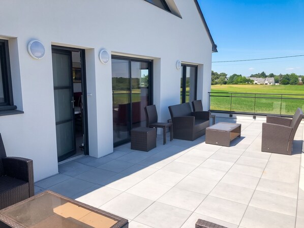 A modern outdoor patio with wicker furniture, a glass door entrance, and a view of a green field.