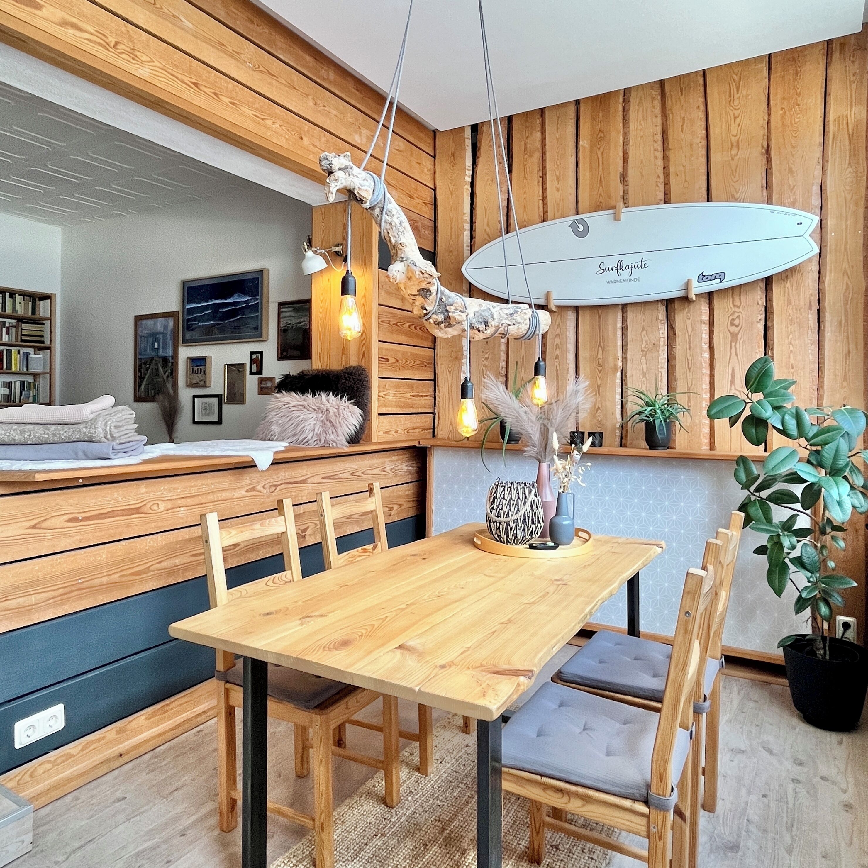 A modern dining area with a wooden table and chairs, a surfboard decoration on the wall, and a bookshelf filled with books.
