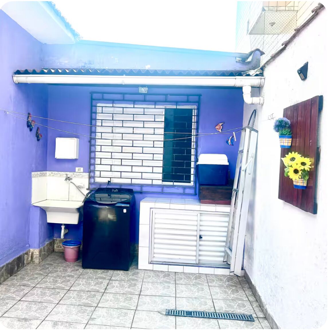 Un petit espace extérieur clos, avec un mur bleu, une fenêtre dotée d’une grille métallique, un évier blanc, une poubelle noire et une porte en grille métallique.