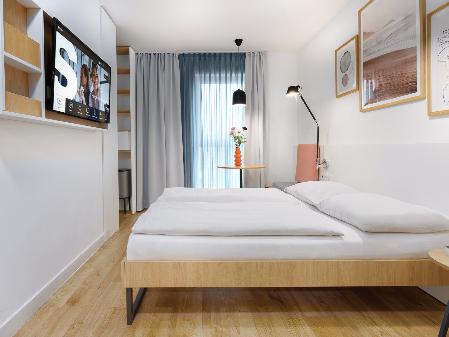 A modern bedroom with a bed, a bedside table with a lamp, a small table with a vase of flowers, and a TV mounted on the wall.