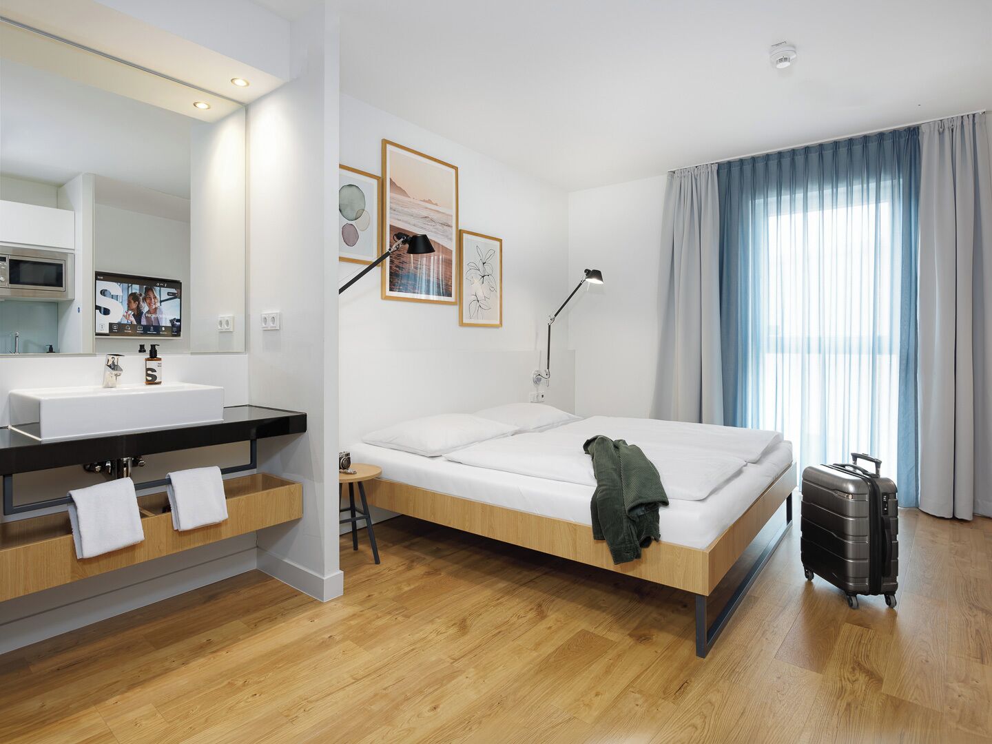 A hotel room with a bed, a bedside table, a mirror, a sink, and a window with curtains.