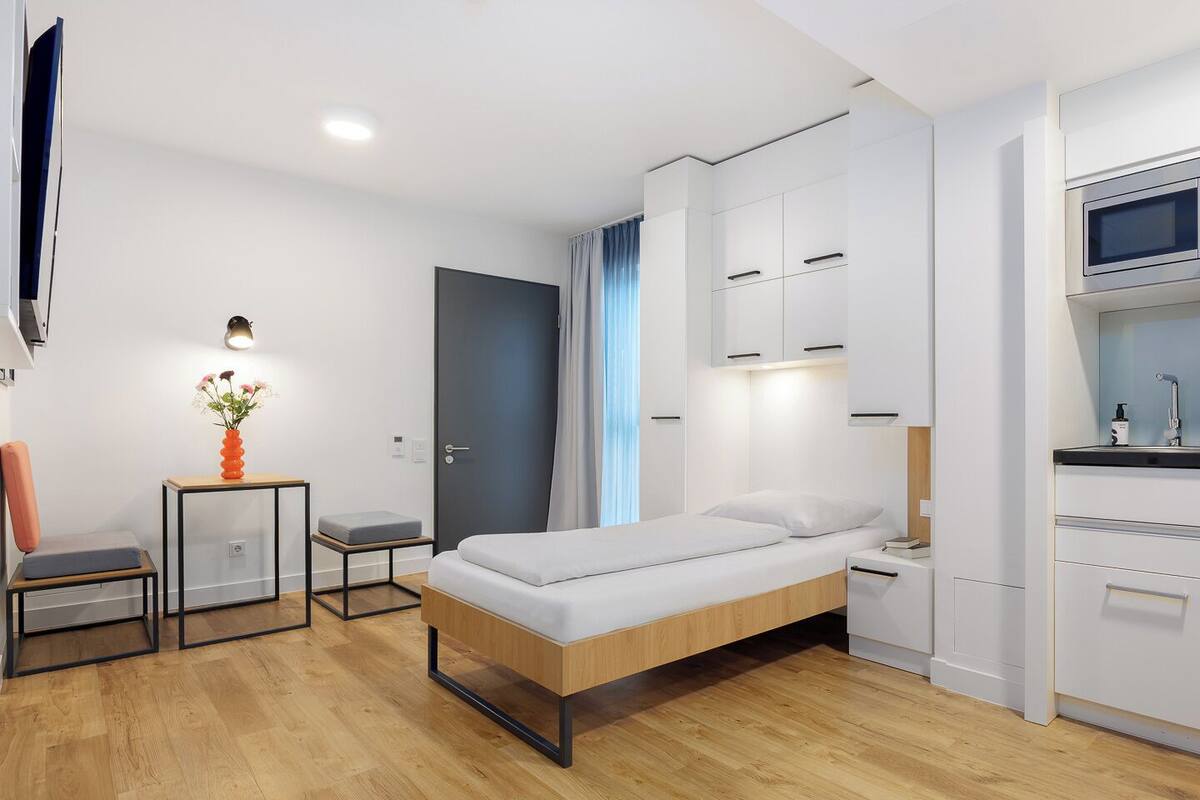 Une chambre d’hôtel moderne équipée d’un lit, d’un petit bureau, d’une chaise, d’une télévision et d’une kitchenette.