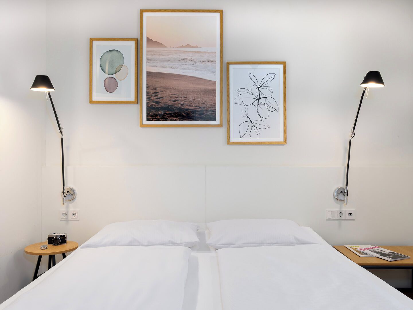 Une chambre d’hôtel avec un lit, deux lampes de chevet, un appareil photo posé sur une petite table et trois œuvres d’art encadrées accrochées au mur.