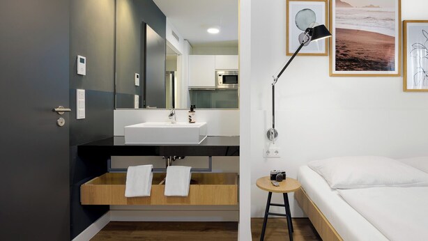 Een moderne badkamer met een grote spiegel, een wastafel en een meubel met handdoeken. Er staat een bed met een wit matras en kussens, en een klein rond tafeltje met een lamp.