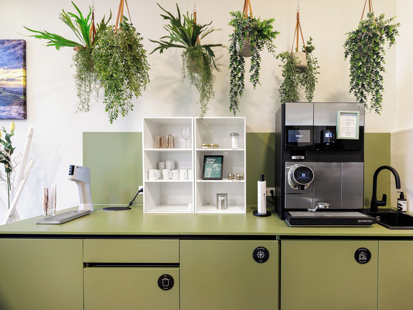 A modern kitchen with a built-in espresso machine, a coffee machine, and a sink. There are hanging plants above the counter and a white shelving unit with kitchenware.