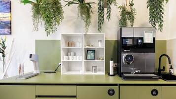 A modern kitchen with a built-in espresso machine, a coffee machine, and a sink. There are hanging plants above the counter and a white shelving unit with kitchenware.