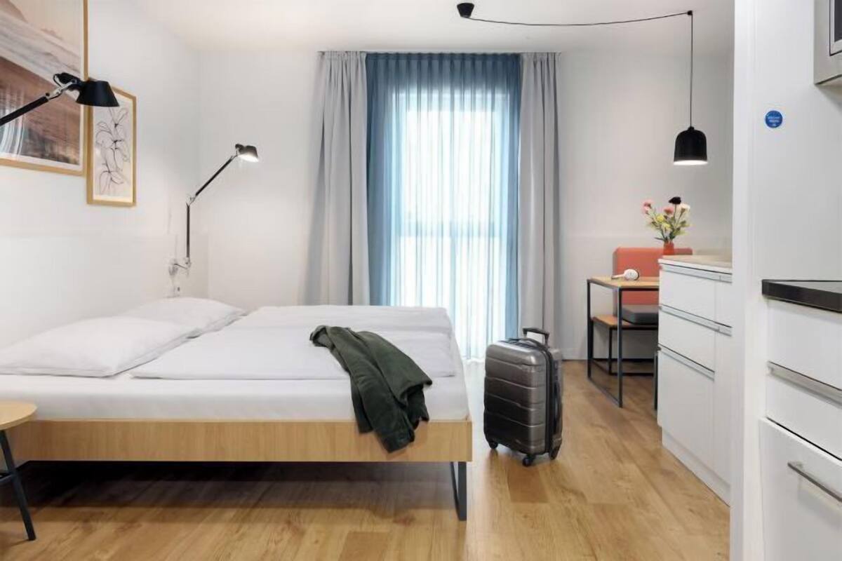 Une chambre d’hôtel avec un lit, un bureau, une chaise, une valise, une lampe et une fenêtre avec des rideaux.