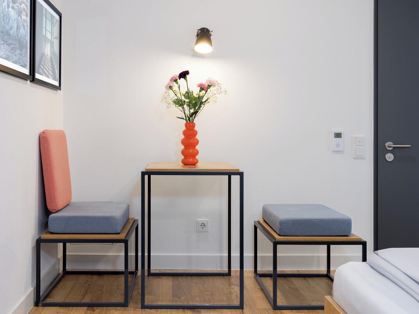 Una habitación pequeña y moderna con dos zonas de estar azules con cojines a juego, una mesa de madera con un florero y una lámpara de pared, además de una puerta con cerradura digital.