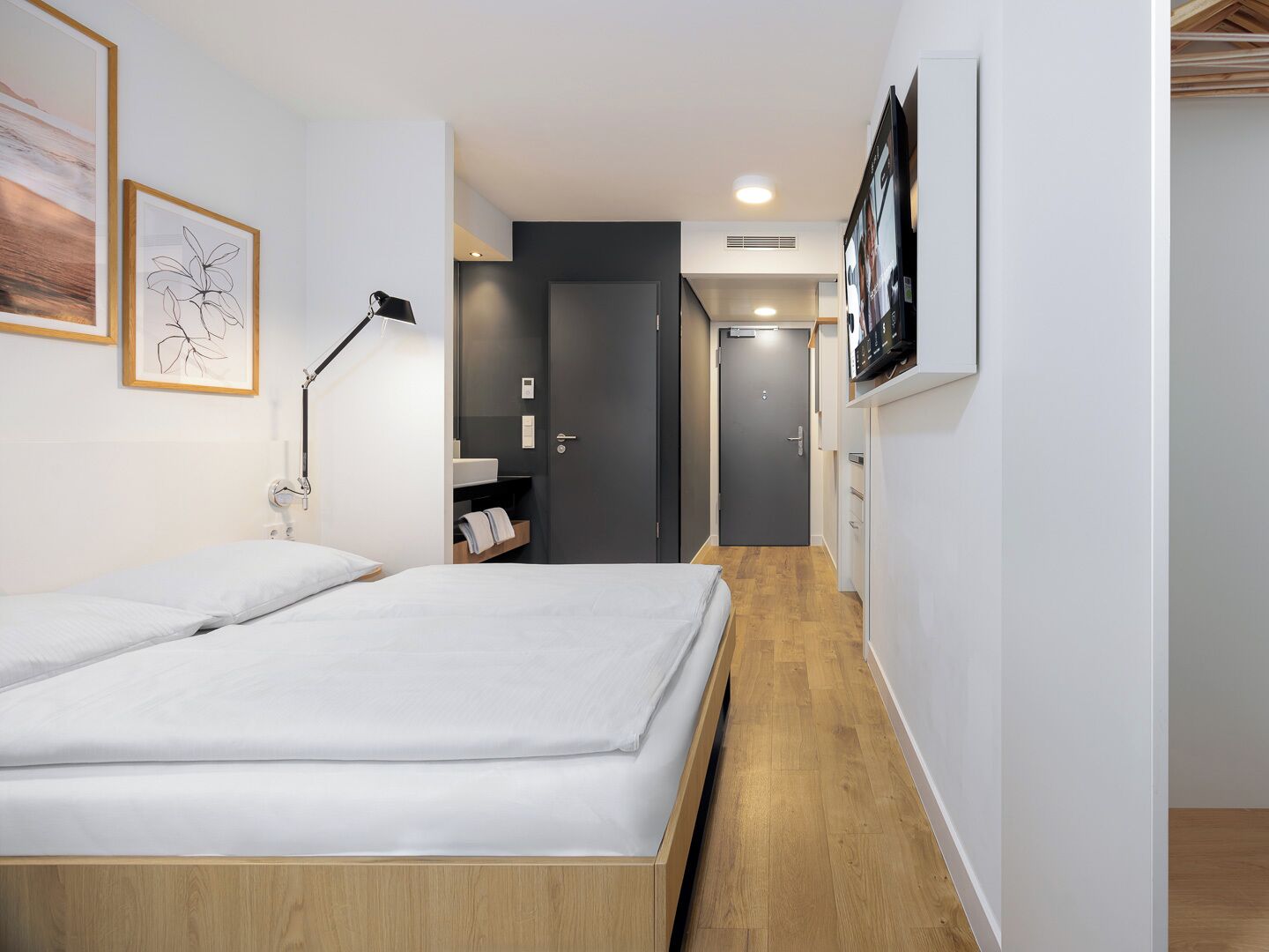 Ein modernes Hotelzimmer mit einem großen Bett, einer Nachttischlampe und Holzboden. Es gibt eine kleine Ablagefläche mit Handtüchern und ein gerahmtes Bild an der Wand.