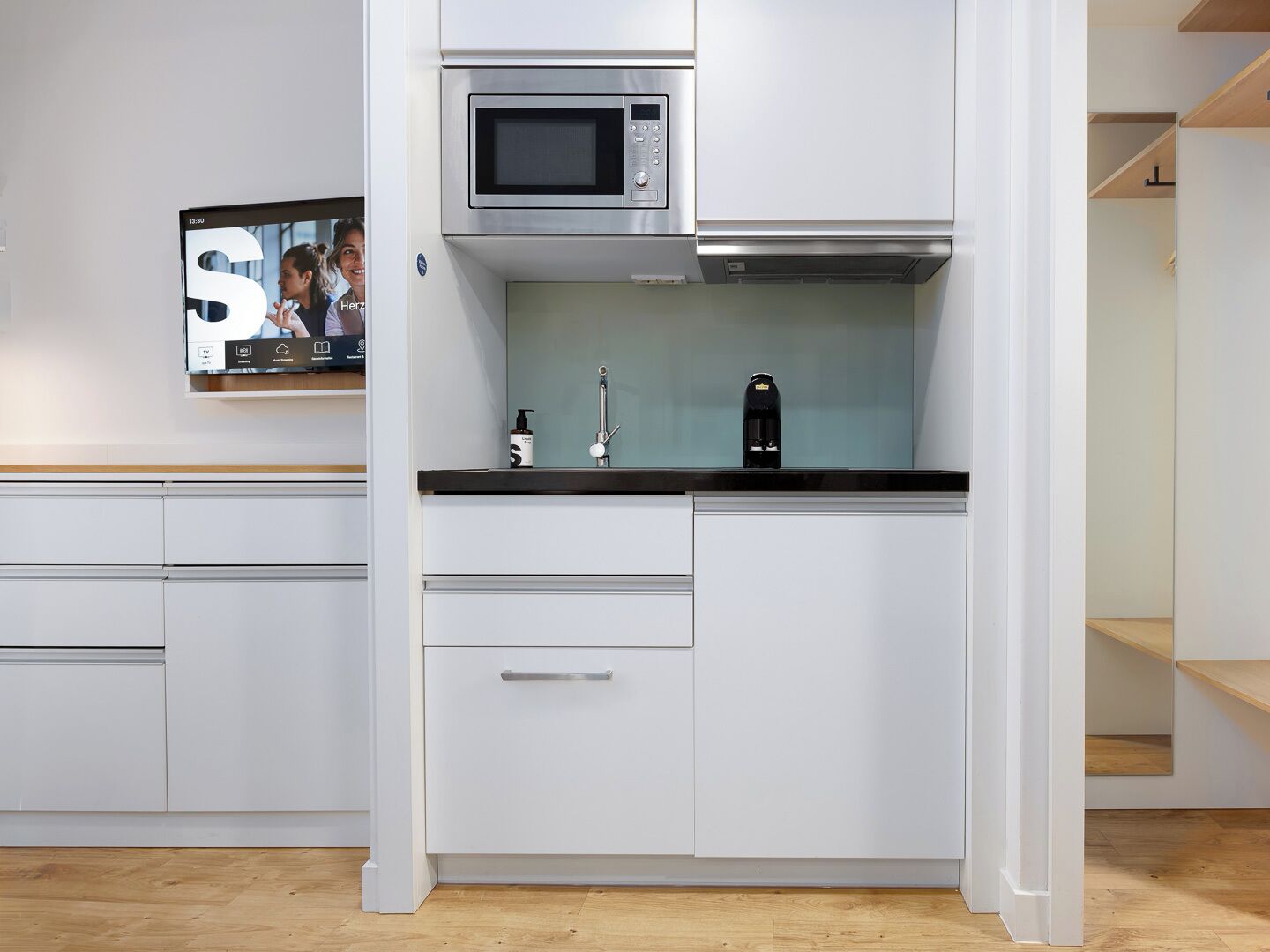 Una cocina moderna con armarios blancos, un microondas de acero inoxidable, una cafetera y un televisor montado en la pared.