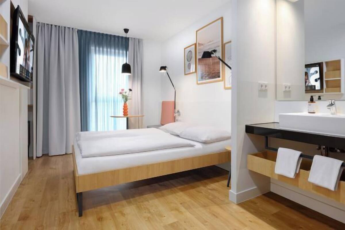 Une chambre d’hôtel moderne avec un plancher en bois, un lit recouvert de draps blancs, une table de chevet avec une lampe, et une salle de bain équipée d’un lavabo et d’un porte-serviettes.