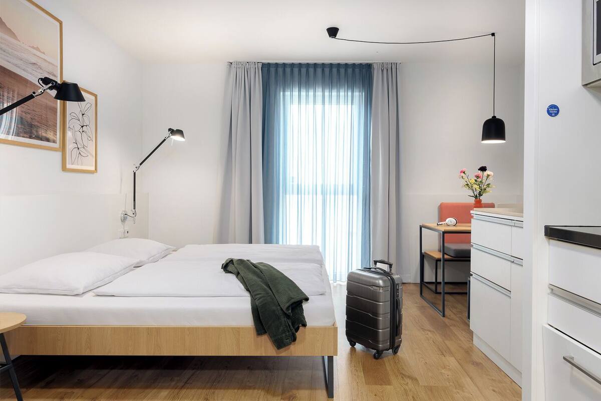 Une chambre d’hôtel moderne équipée d’un lit, d’une table de chevet, d’une lampe, d’une valise, d’une kitchenette et d’une fenêtre avec des rideaux.
