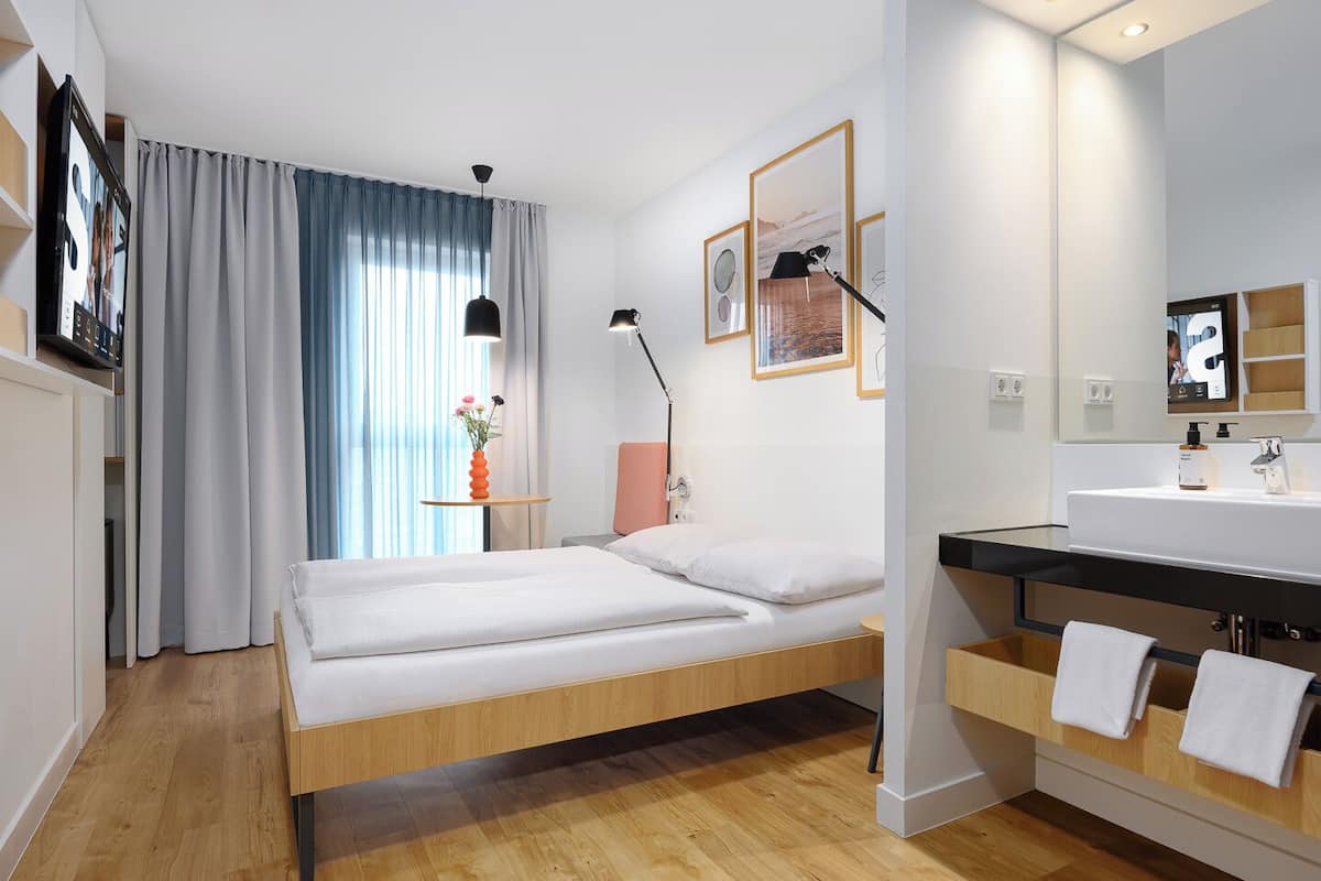 Une chambre d’hôtel moderne avec un lit en bois, une table de chevet avec une lampe, une petite table agrémentée de fleurs, une grande fenêtre avec des rideaux et une salle de bain équipée d’un lavabo et d’un porte-serviettes.