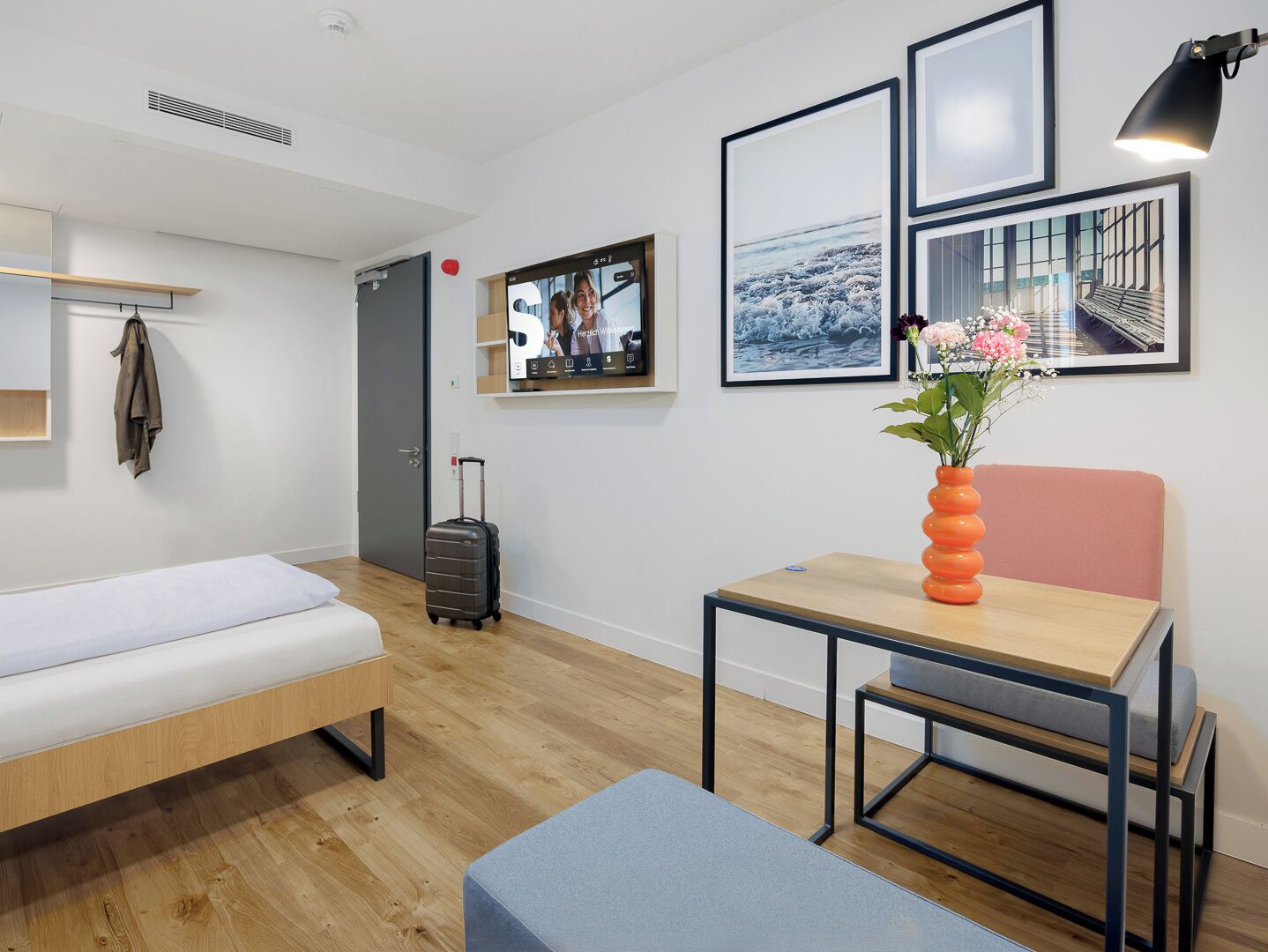 A modern hotel room with a bed, a desk, a chair, a small table, a suitcase, and a wall-mounted TV.