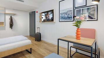 A modern hotel room with a bed, a desk, a chair, a small table, a suitcase, and a wall-mounted TV.