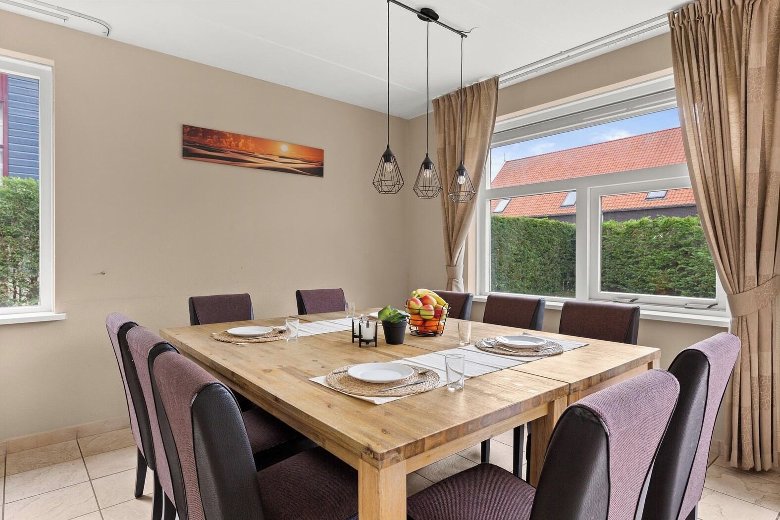 Een eetkamer met een houten tafel voor zes personen, omringd door stoelen, met uitzicht op een gebouw met rode daken.