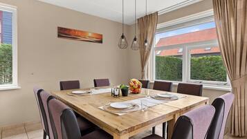 Een eetkamer met een houten tafel voor zes personen, omringd door stoelen, met uitzicht op een gebouw met rode daken.