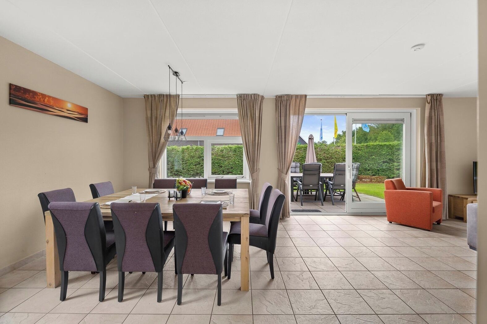Ein Essbereich mit großem Tisch und Stühlen, einem Fernseher und Blick auf einen Garten durch Glastüren.