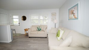 Een woonkamer met een beige bank, een klein rond tafeltje en een rieten fauteuil. Er is een raam met zonwering, een deur en een ingelijst schilderij aan de muur.