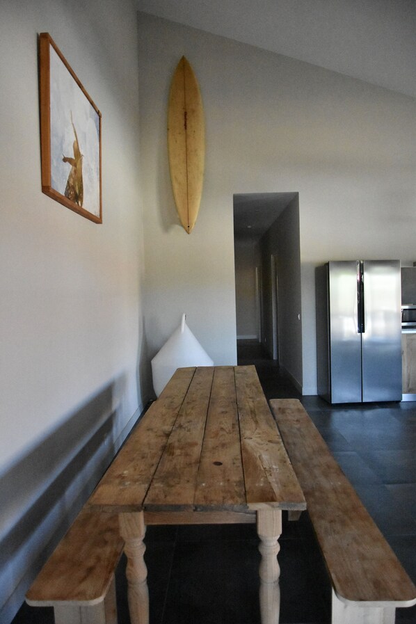 Una mesa de comedor de madera con bancos, una tabla de surf en la pared, un refrigerador y un cuadro enmarcado.