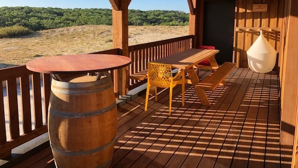 Una terraza de madera con una mesa, sillas, un barril de madera y un colgador con luz.