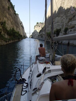A narrow waterway with rocky cliffs on one side and a sailboat with two people on deck. - Sailing Yacht by Owner, Holidays to Greek Islands (Alimos)
