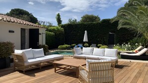 Una terraza de madera con mobiliario de mimbre, piscina y abundante vegetación.