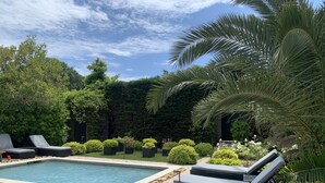 Una piscina con sillas de descanso y un borde de piedra, rodeada de follaje y un seto.