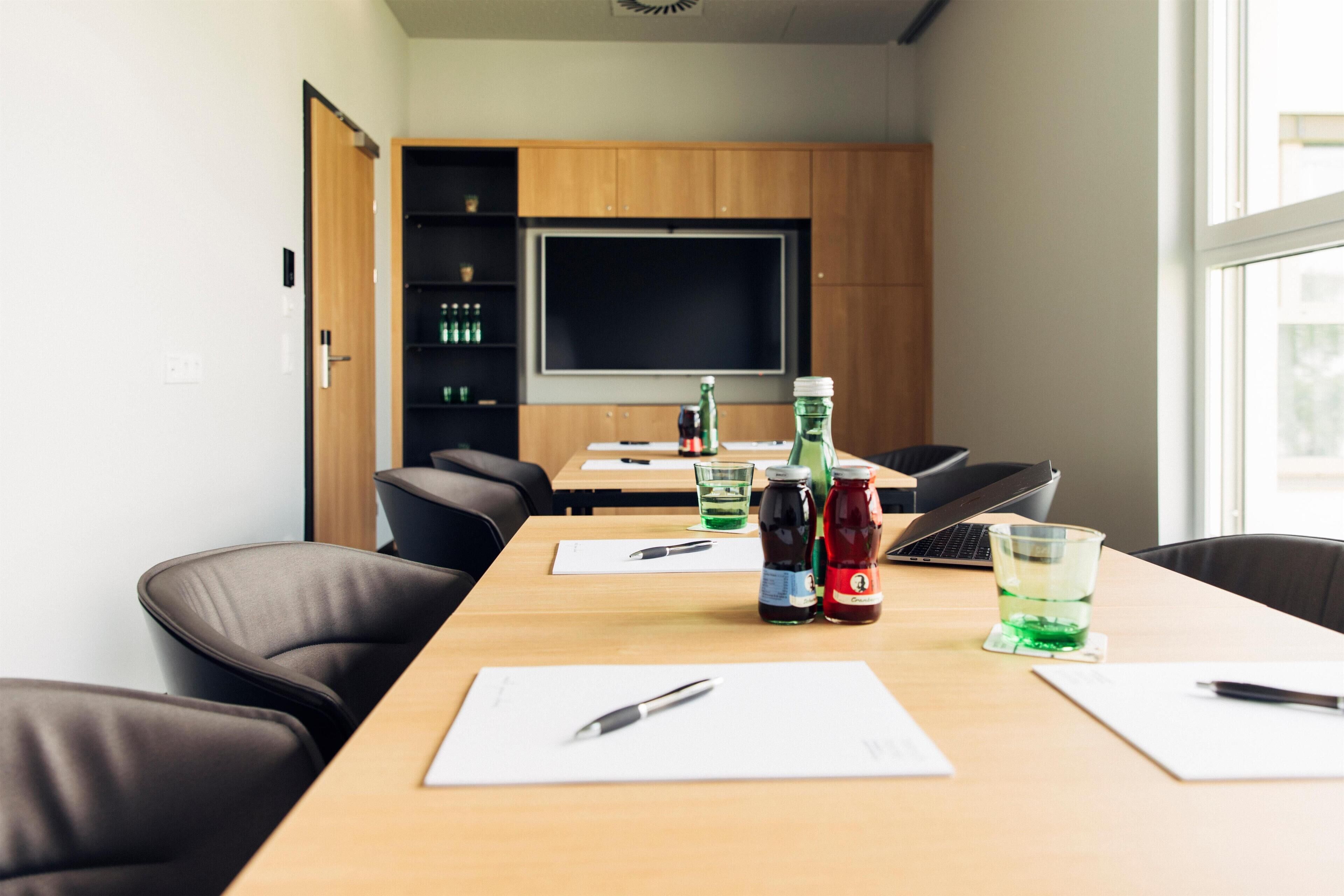 A conference room with a long table, chairs, a projector screen, and a shelf with bottled beverages.