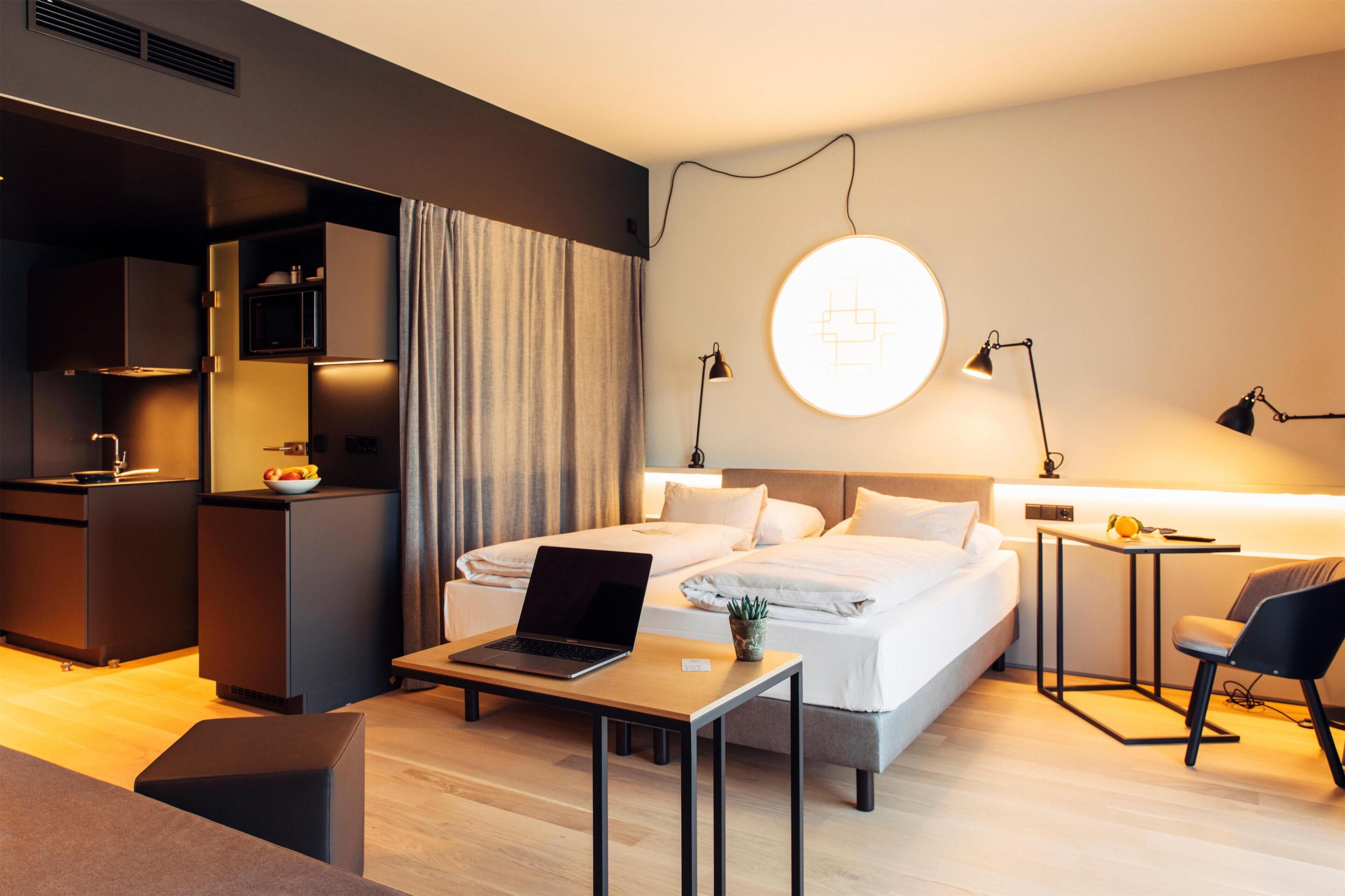A modern hotel room with a bed, a desk with a laptop, a small table, a chair, a bedside table with a lamp, and a wall-mounted light fixture.