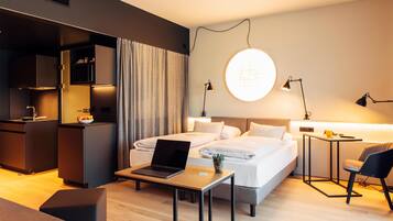 A modern hotel room with a bed, a desk with a laptop, a small table, a chair, a bedside table with a lamp, and a wall-mounted light fixture.