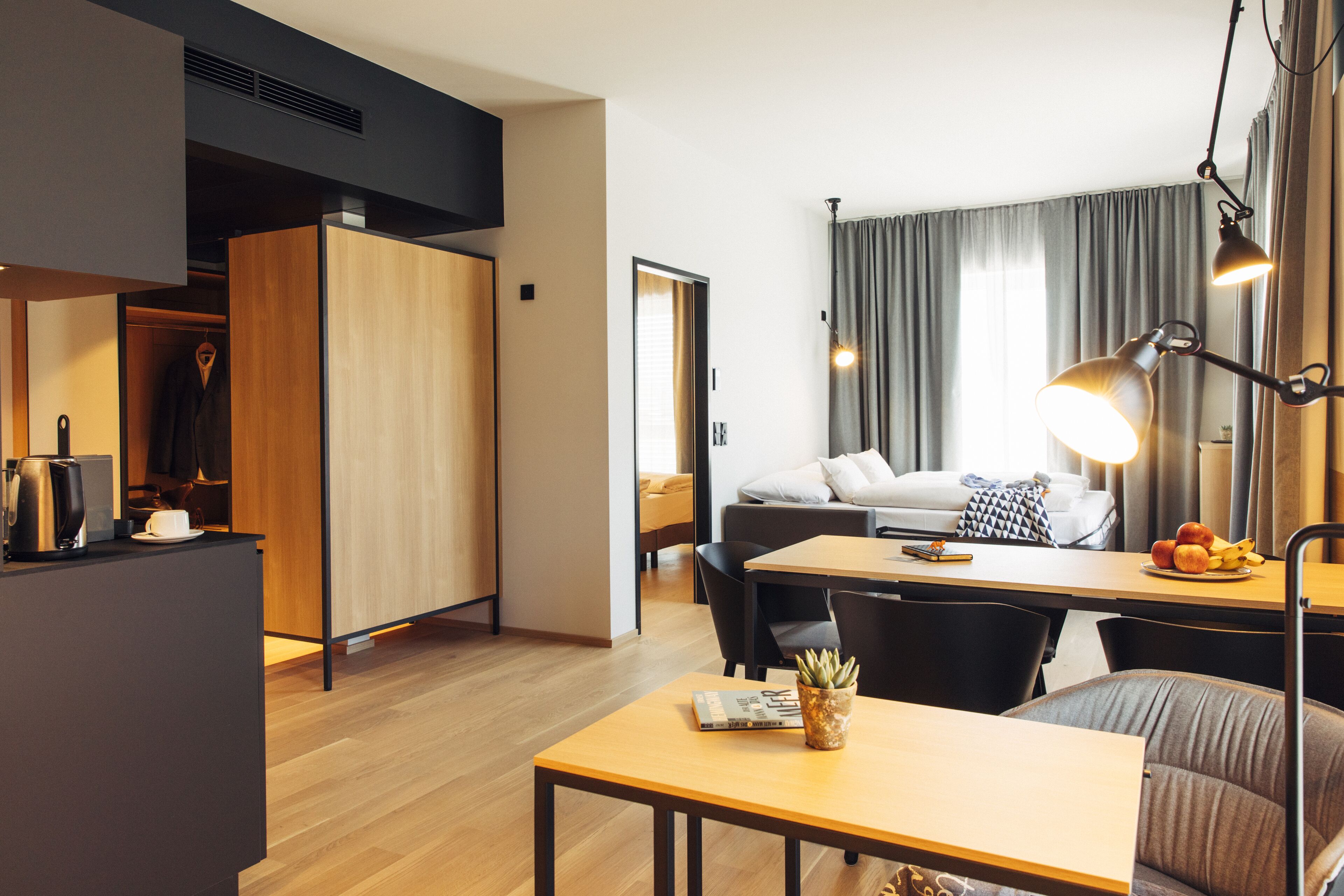 A modern hotel room with a wooden dining table, a sofa, and a desk with a lamp.