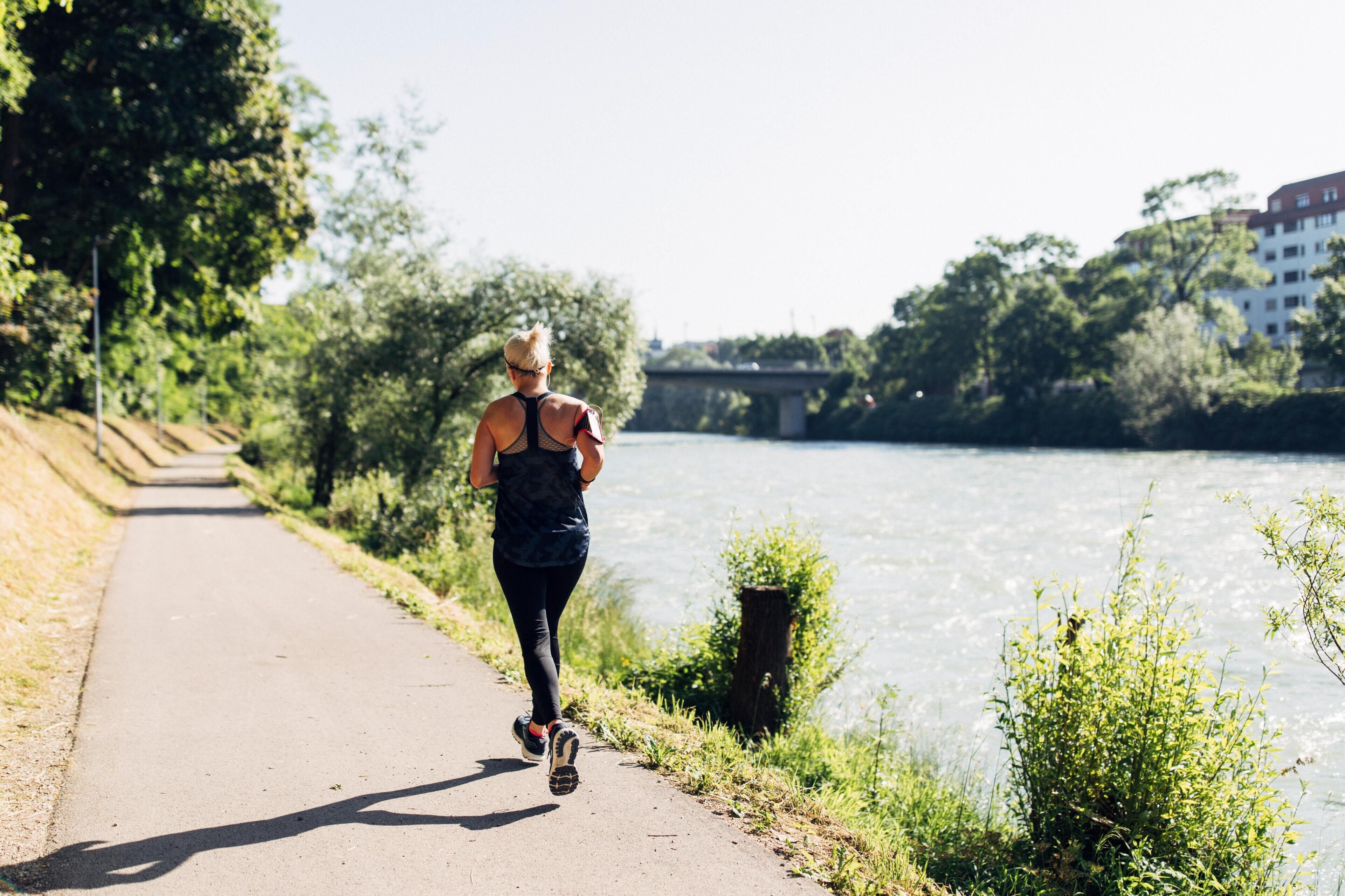 A person jogging on a paved path beside a river, with trees and a bridge in the background.
