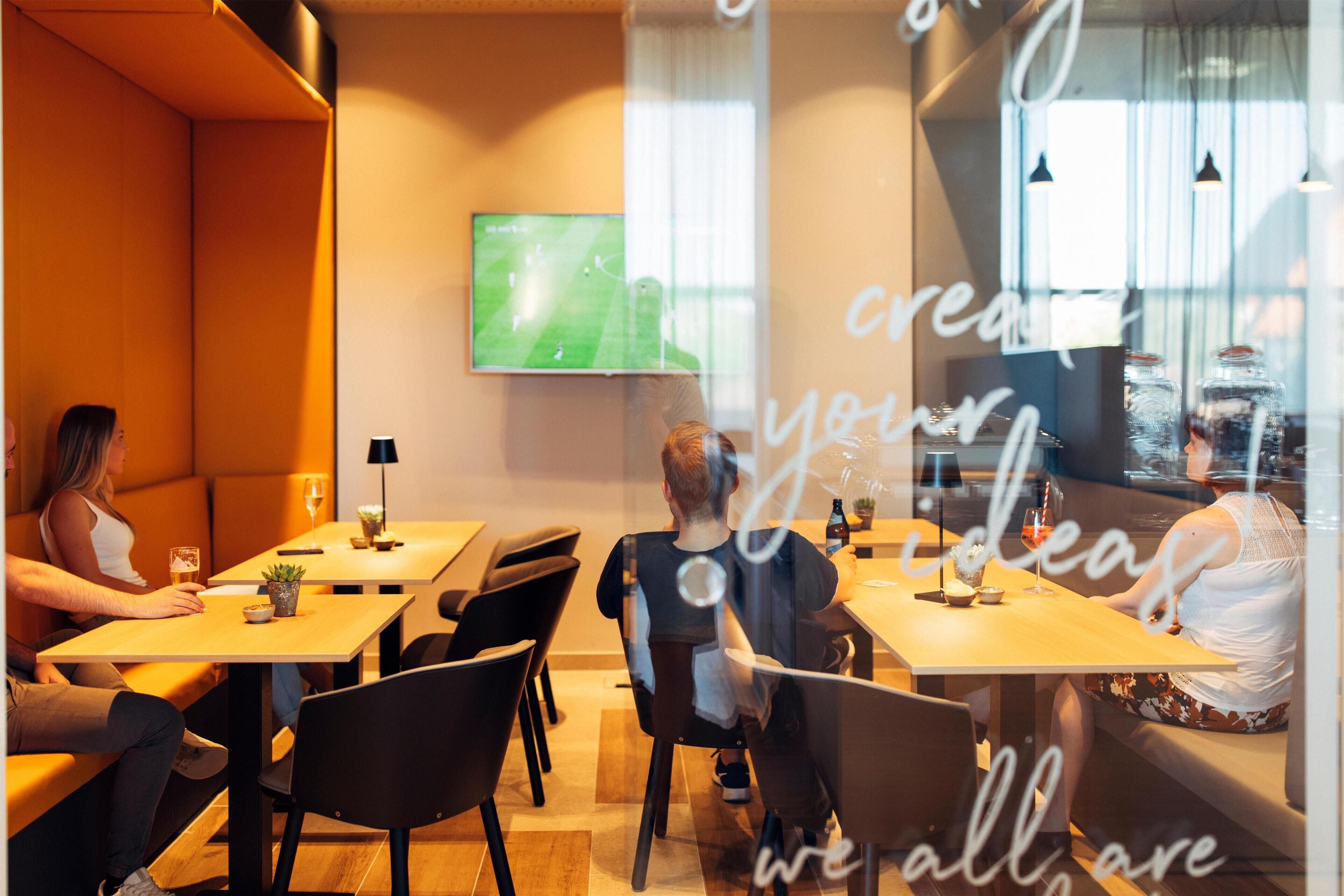 A restaurant with tables and chairs, a TV screen displaying a soccer game, and a glass wall with text.
