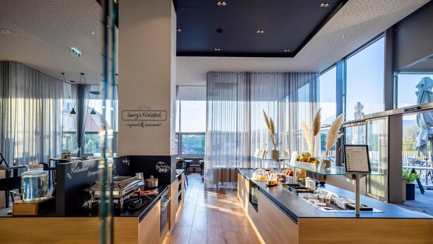 A modern cafe with a wooden counter, a menu board, and a view of the city through large windows. - harry’s home Steyr hotel & apartments (Steyr)