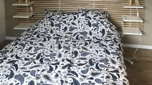 Une chambre à coucher comprenant un lit, une tête de lit, deux tables de chevet avec des lampes, une œuvre d’art accrochée au mur et une étagère près du lit.
