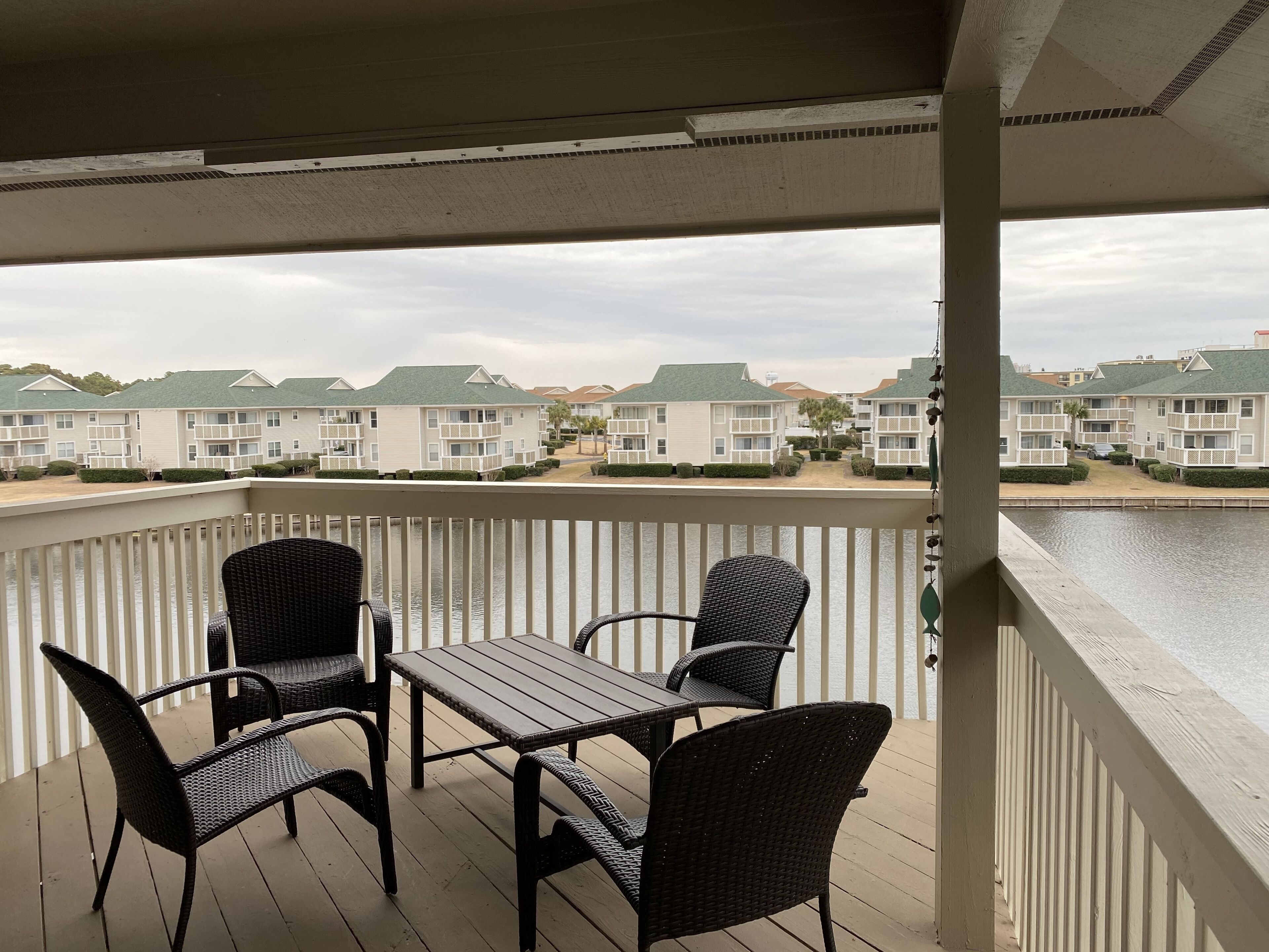 A balcony with wicker chairs and a table, overlooking a marina with several white buildings with green roofs.