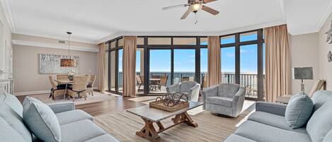 A spacious living room with a large window, a ceiling fan, and a dining area with a view of the ocean.