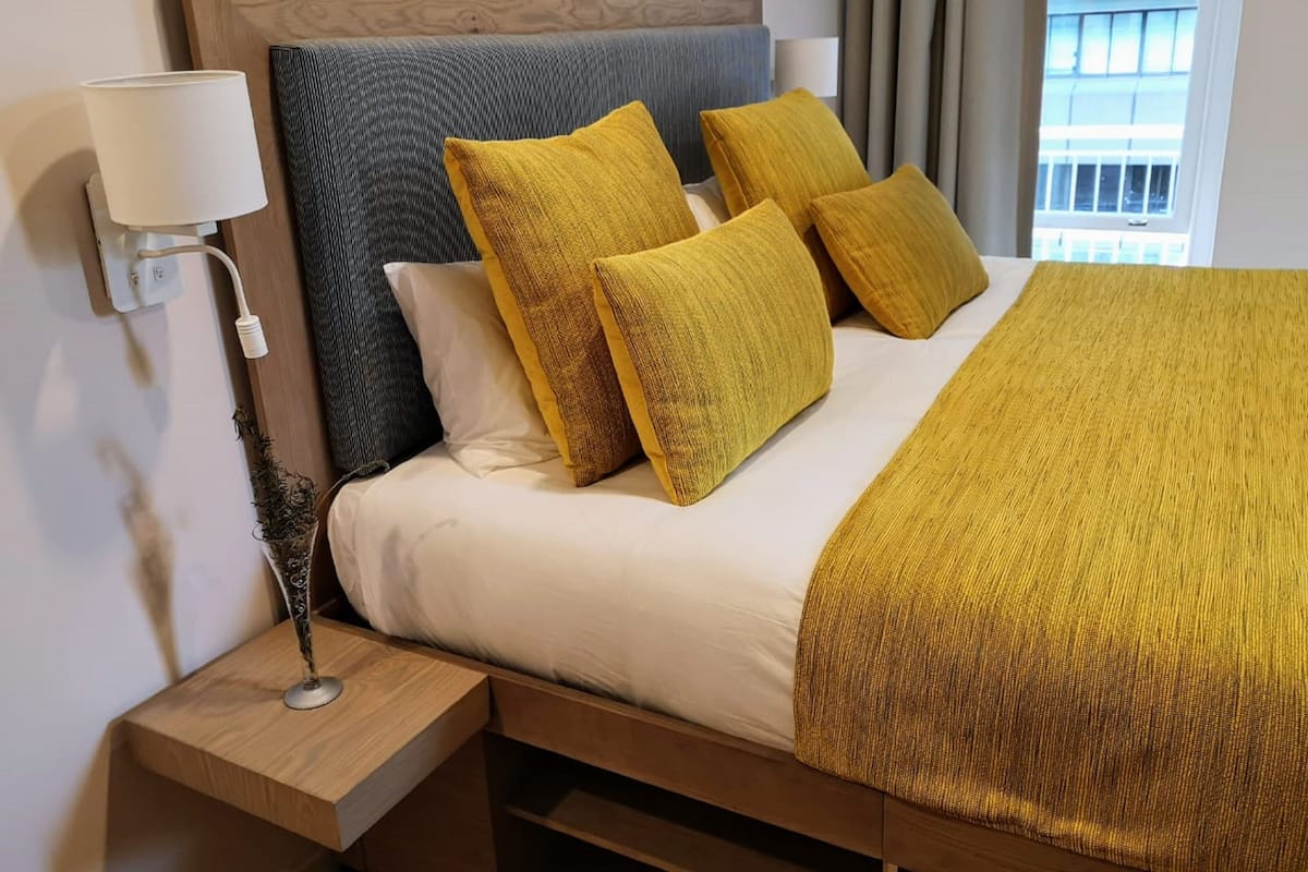 Una cama bien hecha con un edredón amarillo, una mesita de noche de madera con una lámpara y vistas al exterior a través de una ventana.