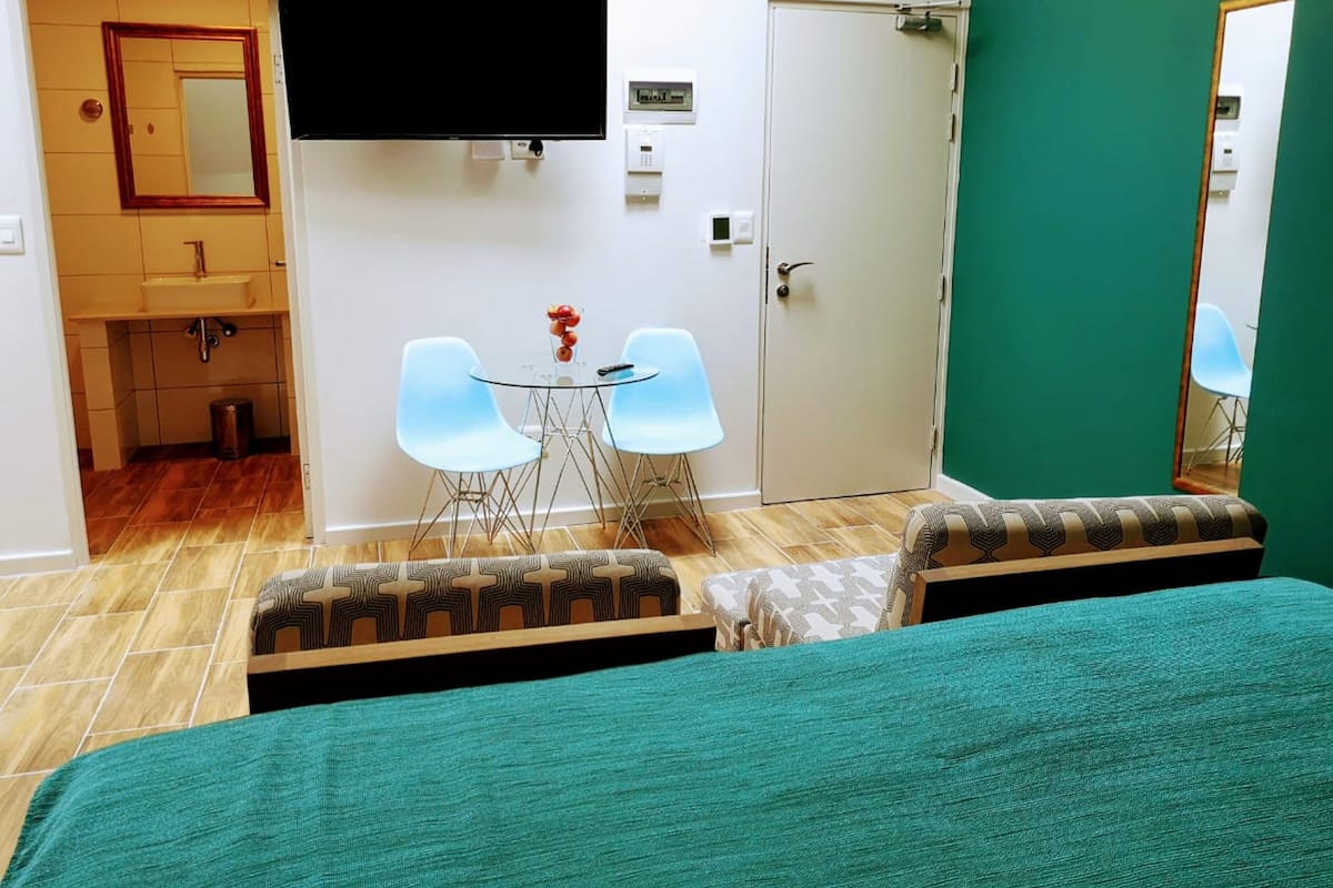 Habitación de hotel con cama, televisor, una mesita con sillas, baño y pared verde.