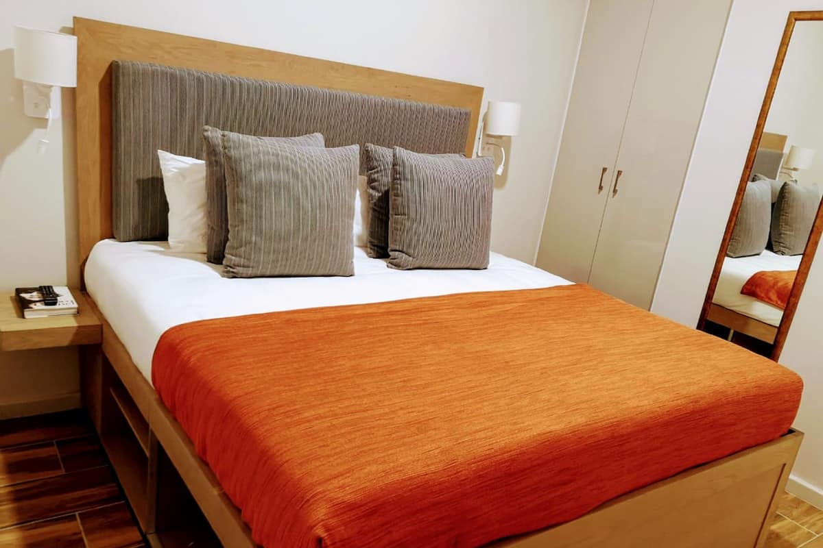 Una cama bien hecha con cabecera de madera, ropa de cama blanca y una manta roja. Hay dos mesitas de noche con lámparas y un espejo en la pared.