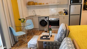 Una kitchenette compacta con lavadora, horno y microondas. Hay un pequeño comedor con una mesa de vidrio y dos sillas. Un sofá con funda estampada se encuentra en la zona de estar.