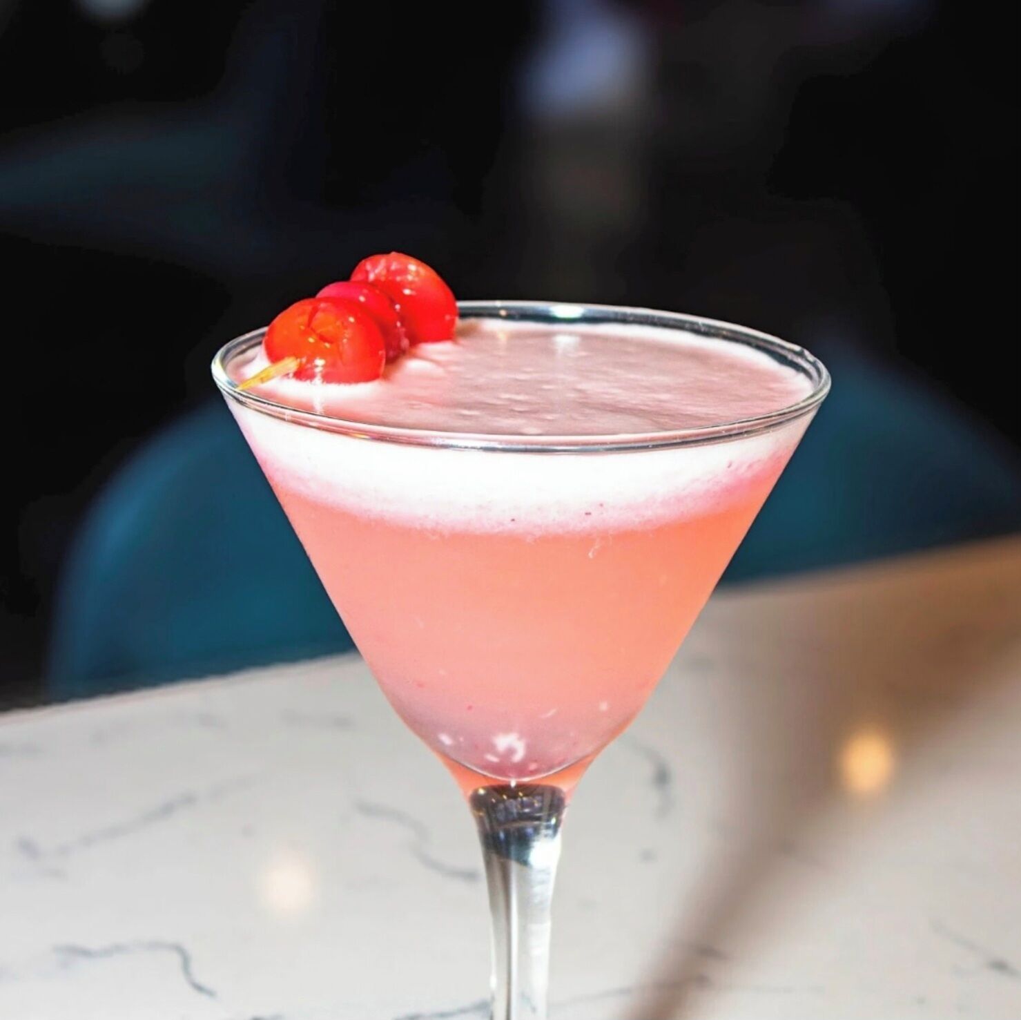 A cocktail in a martini glass with a pink hue and a cherry garnish.