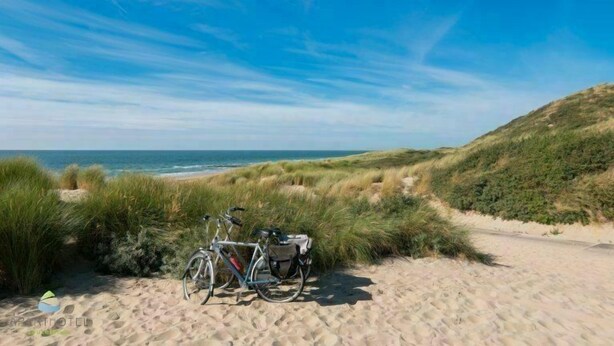 Una bicicleta estacionada en un sendero arenoso con dunas y una playa al fondo.
