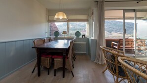 A dining area with a wooden table and wicker chairs, a hanging light fixture, and a view of a mountainous landscape through large windows. - Apt - Duinweg 117-4 | Zoutelande 'Duinoo (Zoutelande)