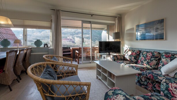 A spacious living room with a large window, a sofa set, a television, and a dining area with wicker chairs. - Apt - Duinweg 117-4 | Zoutelande 'Duinoo (Zoutelande)