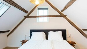 A bedroom with a sloped ceiling, a bed with white bedding, a small window, and a hanging light fixture.