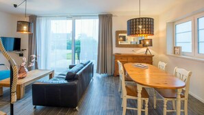 A modern living room with a sofa, dining table, and chairs. There is a TV mounted on the wall and a large window providing natural light.