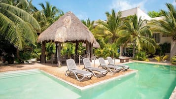 A pool area with lounge chairs, a thatched-roof structure, and palm trees.