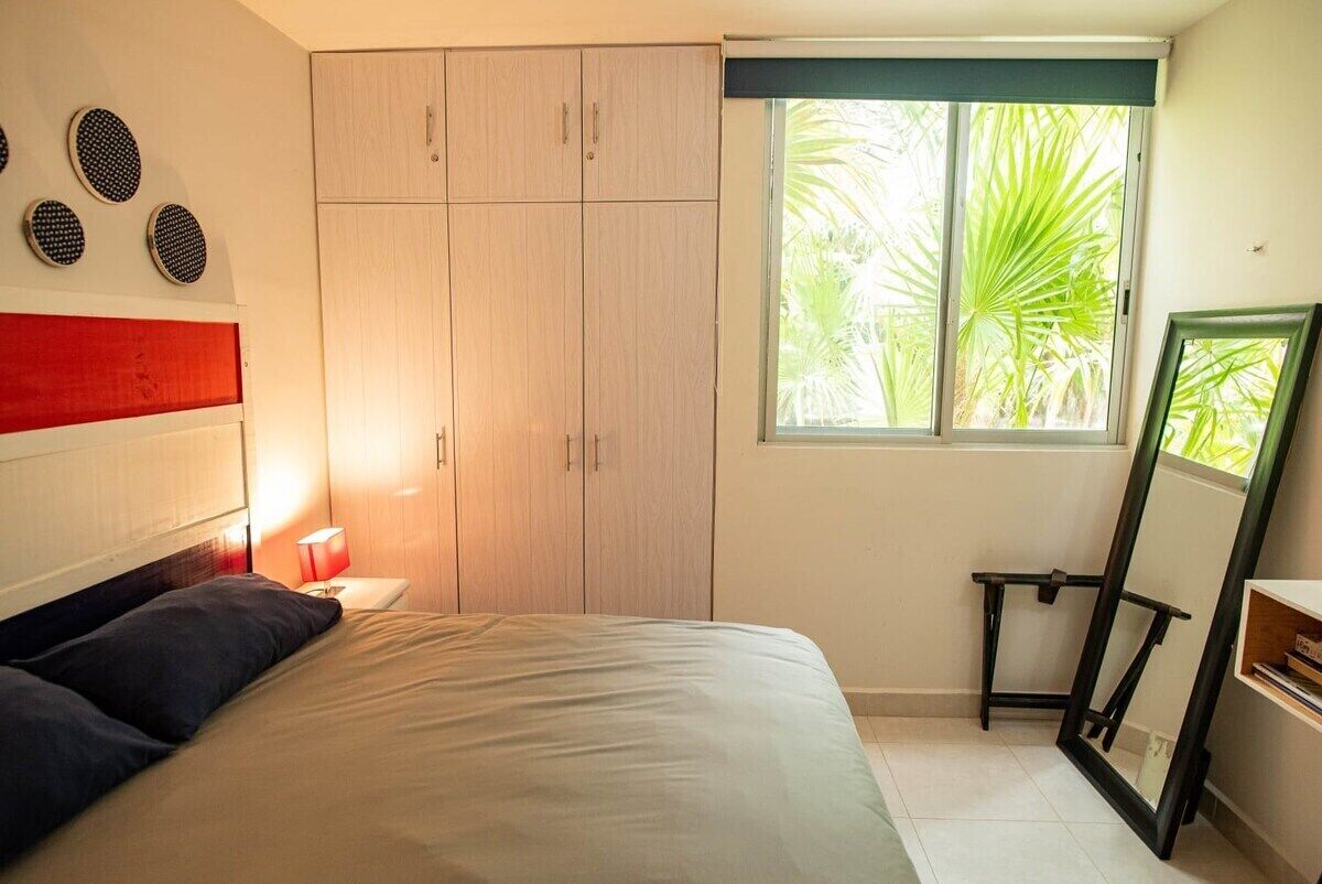 A bedroom with a bed, a wardrobe, a window with a view of palm trees, and a mirror.